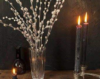 A clear glass vase filled with white pussywillow stems, styled on a dark wooden surface next to two lit black taper candles and a small amber bottle against a dark textured wall. sold by The Stick And Stem