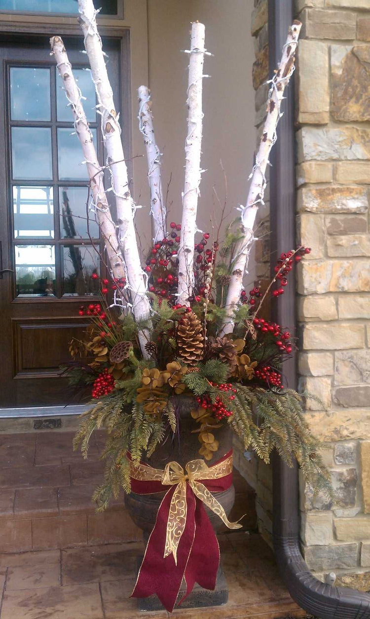 A festive outdoor holiday plant arrangement in a dark stone pot, featuring white birch logs, red berries, pinecones, green foliage, and a burgundy and gold ribbon bow, set on a front porch.