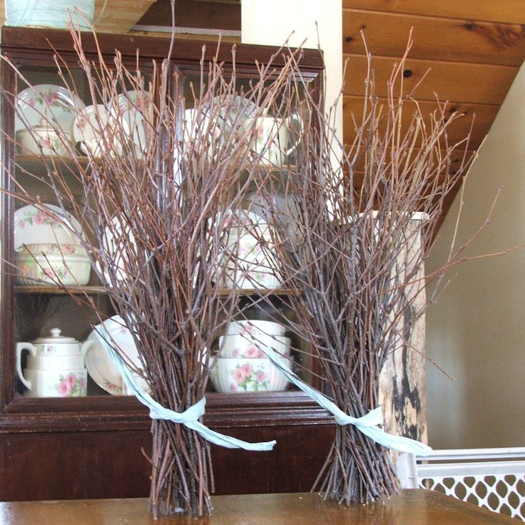 Two bundles of dried brown birch twigs tied with light blue ribbon, displayed on a wooden table in front of a dark wood china cabinet. Sold By The Stick And Stem