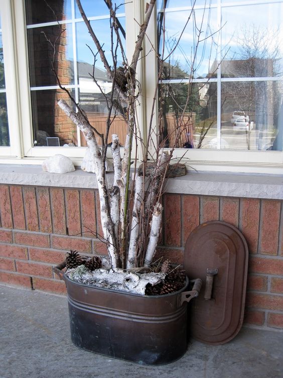 A rustic decorative planter made from a dark metal tub with a rusted brown lid leaning against it, filled with white birch logs, pinecones, and white faux snow, set against a brick wall and window.