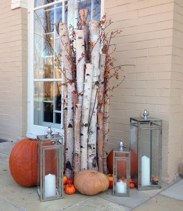 Outdoor fall porch decor arrangement with stacked birch logs, orange pumpkins, small gourds, and two tall glass lanterns with white candles against a beige brick wall.
