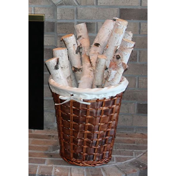 A brown woven basket filled with birch logs, lined with a white fabric liner, set against a brick fireplace background. Sold by The Stick And Stem