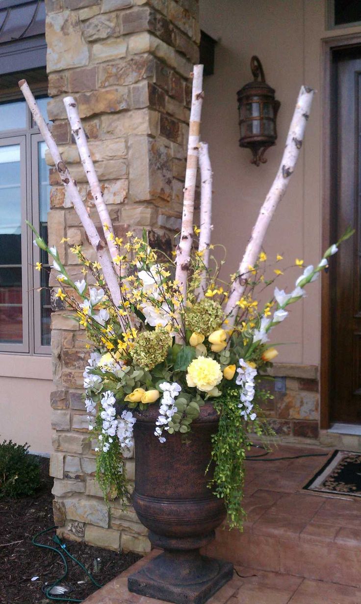 A large rustic dark brown vase filled with a floral arrangement featuring white birch logs, yellow wildflowers, and green foliage, set on a stone porch next to a stone pillar.