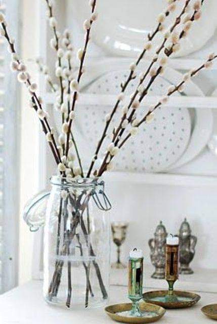 A clear glass jar vase filled with pussywillow branches featuring pale cream buds, styled on a white tabletop with decorative candles and plates in the background.