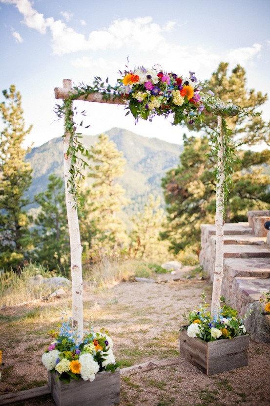 A rustic wooden archway made from birch poles sold by The Stick And Stem decorated with colorful wildflowers and greenery, set outdoors in a mountain landscape with trees and stone steps in the background.