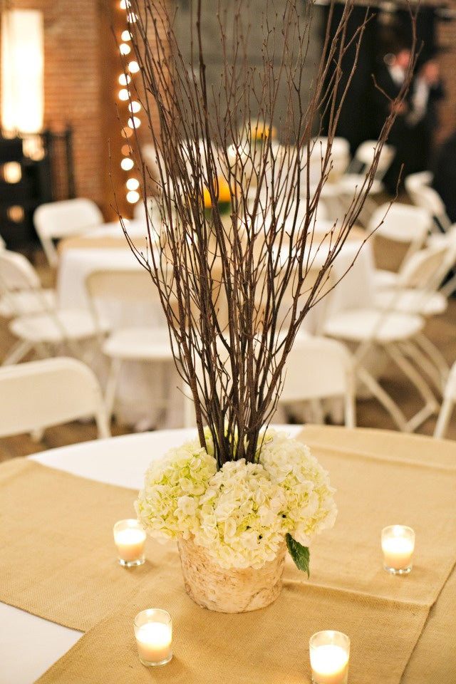 A rustic wedding centerpiece featuring white hydrangeas in a textured beige pot, surrounded by tall brown birch branches, set on a burlap runner with small lit candles at a reception table. Sold By The Stick And Stem