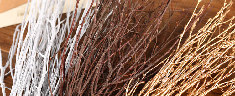 Three bundles of decorative birch twigs or branches in white, brown, and coppery gold, arranged on a wooden surface.