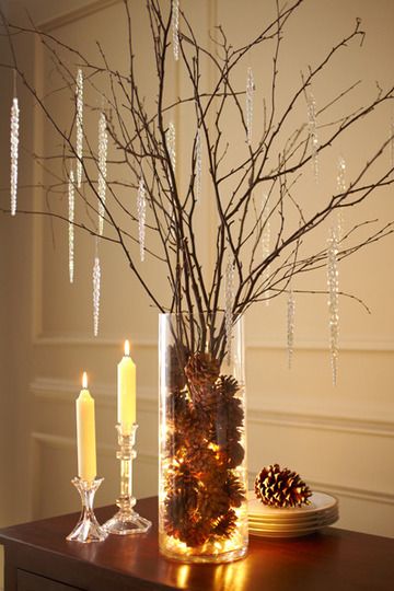 A clear glass vase filled with birch twigs  warm white fairy lights, surrounded by bare winter branches with hanging clear icicle ornaments, displayed on a dark wooden table beside two lit cream candles and a small plate with another pinecone